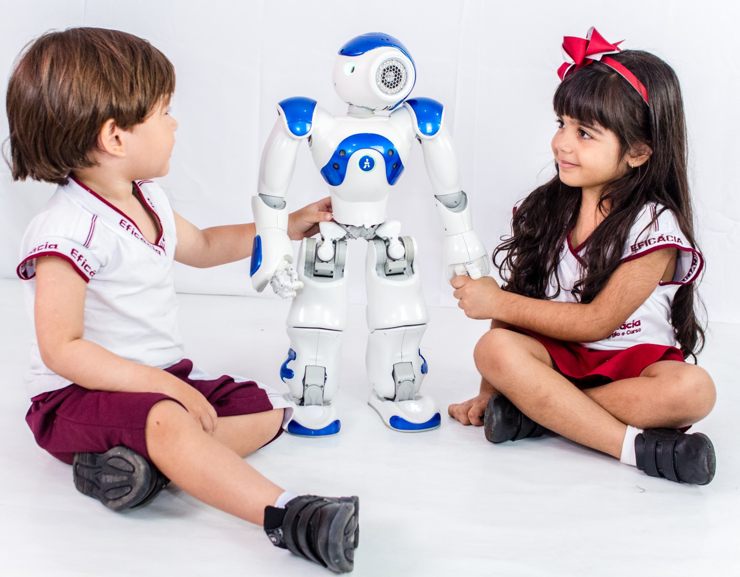 Duas crianças com uniforme do Eficácia (um menino e uma menina) interagem com um robô branco e azul utilizado, normalmente, em aulas de robótica educacional.