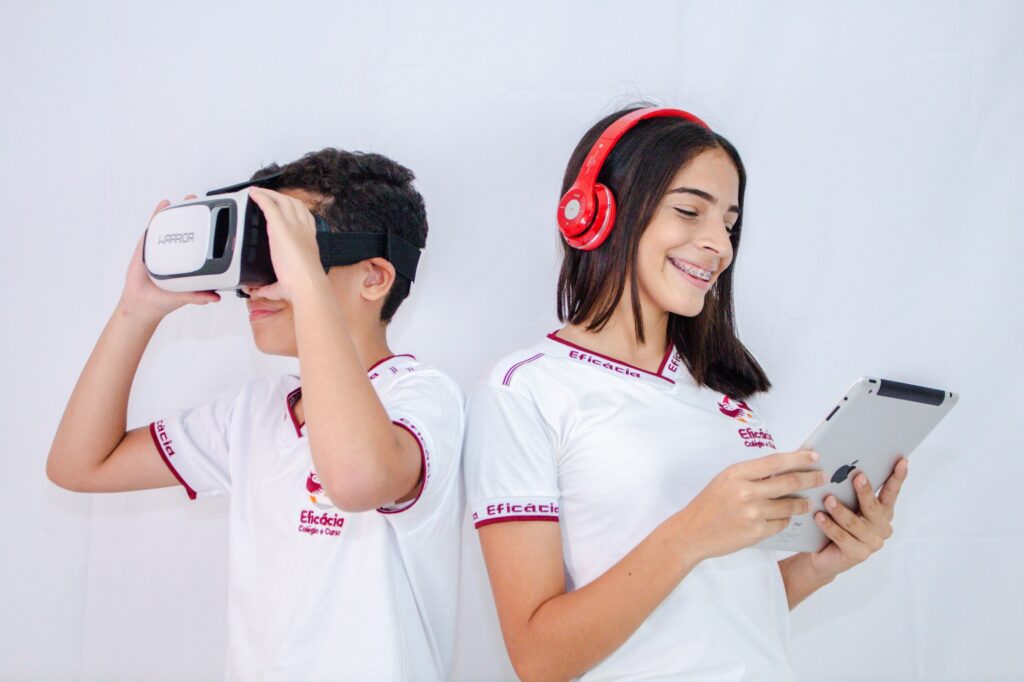 Dois jovens usam uniforme do Eficácia e utilizam aparelhos eletrônicos. Um menino usa um óculos de realidade virtual. Uma menina usa um tablet e headphones vermelhos.